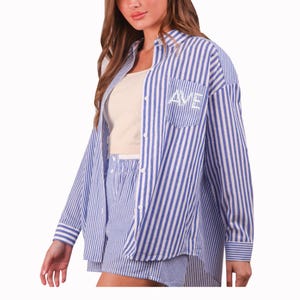 Pode incluir: Um conjunto de pijama listrado azul e branco. O conjunto inclui uma camisa de botão de manga comprida com um bolso no peito e as letras bordadas "AVE", e calções a condizer. A modelo está a usar um top creme por baixo.