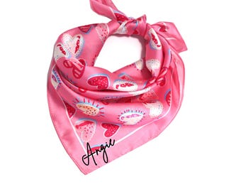 May include: Pink silk bandana with a white border and a pattern of colorful hearts. The bandana is tied in a knot and the name "Angie" is written in black cursive on the bottom corner.