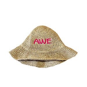 May include: A beige crocheted bucket hat with a pink embroidered word "AWE" on the front.