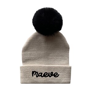 May include: A beige knitted beanie with a black pom pom and the name "maeve" embroidered in black on the front.