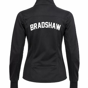 May include: A black long-sleeve jacket with a high collar and a horizontal zippered pocket at the lower back. The word BRADSHAW is printed in large, white, block-style capital letters across the upper back section of the garment.