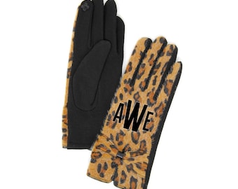 Puede incluir: Un par de guantes de leopardo marrón y negro con un lazo negro y las letras "AWE" bordadas en el dorso de la mano.