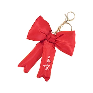 Pode incluir: Chaveiro de laço vermelho com o nome Angie escrito em branco no laço.
