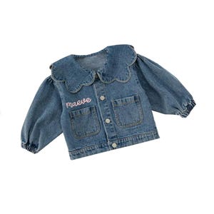 May include: A blue denim jacket with a scalloped collar and a pink embroidered name "maeve" on the front.