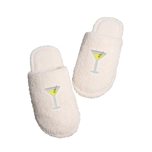 May include: A pair of off-white, plush slippers. Each slipper features an embroidered martini glass with a yellow drink, olive, and a cocktail stick. The slippers are designed for indoor wear and have a soft, textured appearance.