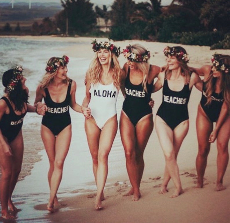 May include: Six women wearing black and white one-piece swimsuits with the words "Aloha" and "Beaches" printed on them. They are standing on a sandy beach with the ocean in the background.