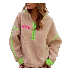 May include: A tan fleece pullover with neon green accents. The pullover features a half-zip closure, a hood, and the word "AWE" in pink letters. The sleeves and sides have neon green stripes.