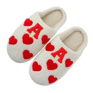 May include: A pair of off-white, plush slippers with red heart accents. Each slipper features a large red letter "A" with gold trim. The slippers are designed for indoor wear and offer comfort and style.