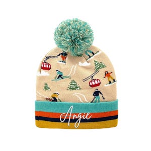 Puede incluir: Un gorro de punto beige con un pompón verde azulado y un patrón de esquiadores, practicantes de snowboard y telecabinas rojos. El puño es verde azulado, naranja, azul marino y amarillo con el nombre Angie en cursiva blanca.