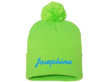 Può includere: Un berretto verde brillante con un pompon soffice in cima. Il berretto ha il nome "Josephine" ricamato sul davanti in blu.