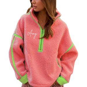 Puede incluir: Un jersey de forro polar color coral con detalles en verde neón. La parte superior tiene media cremallera, capucha y bolsillo delantero. El nombre "Amy" está escrito en blanco. El ribete verde neón está en la cremallera, los puños y las rayas laterales.