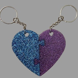 May include: Two glittery heart-shaped keychains, one blue and one purple, designed to fit together like puzzle pieces. Each keychain has a silver keyring and chain. The keychains are on a gray background.