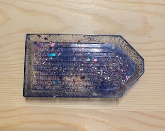May include: A rectangular, dark blue resin tray with a glittery, galaxy-like design. The tray has a series of parallel grooves and a pointed end. The glitter includes silver, pink, and teal colors. The tray is designed for crafting.