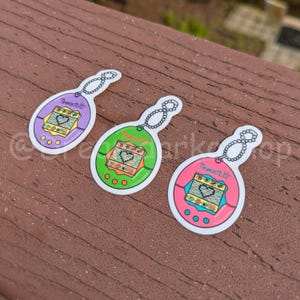 May include: Three colorful stickers in the shape of a classic handheld digital pet game. The stickers are purple, green, and pink, each with a different color scheme. The text "Tamagotchi" is visible on each sticker.