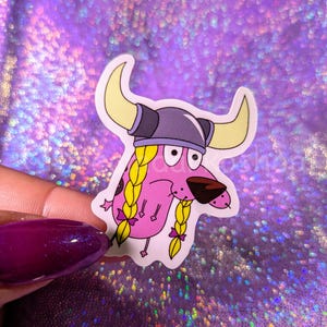 May include: Pink cartoon dog wearing a Viking helmet with blonde braids and pink bows. The dog is looking to the right.