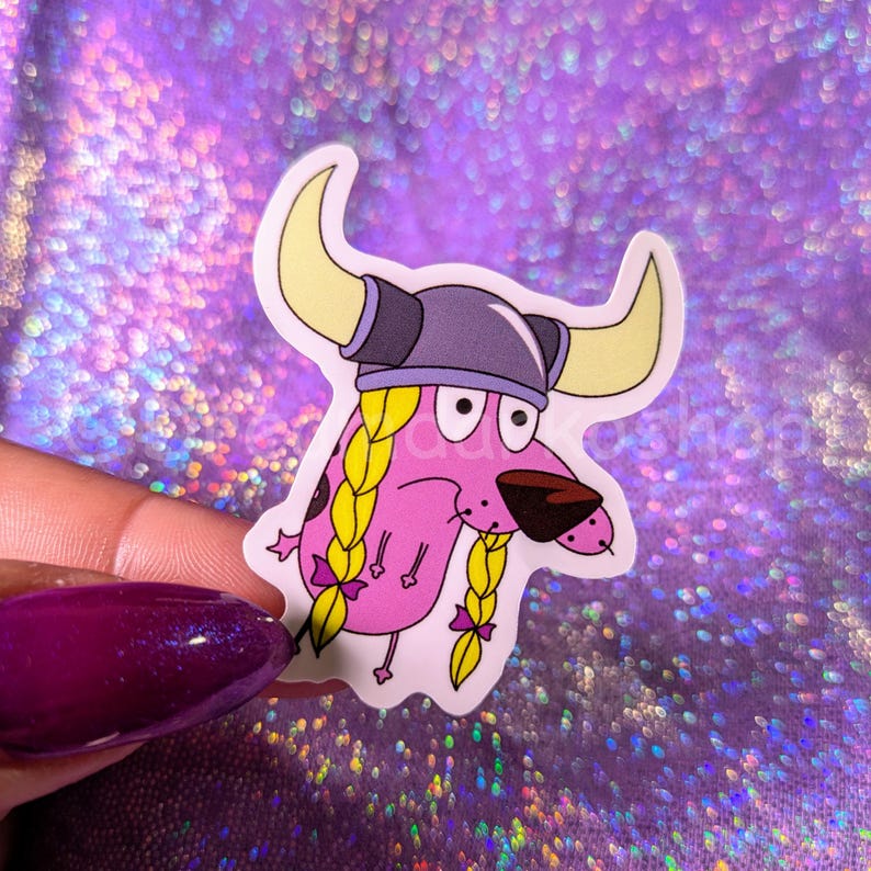 May include: Pink cartoon dog wearing a Viking helmet with blonde braids and pink bows. The dog is looking to the right.