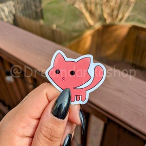 May include: A pink cartoon cat sticker with black eyes and a pink nose. The sticker is being held by a hand with black nail polish.
