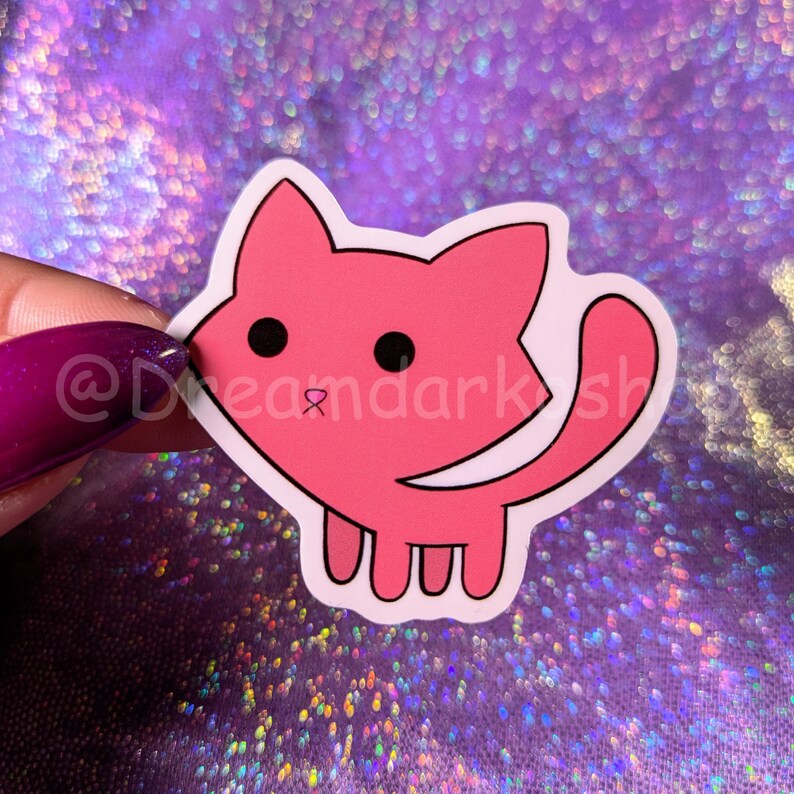 May include: A pink cartoon cat sticker with black eyes and a pink nose. The sticker is on a sparkly purple background.