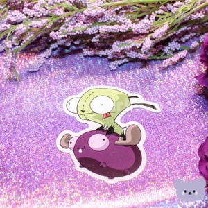 May include: A green cartoon character with a long tongue riding on a purple cartoon creature. The sticker is on a sparkly purple background.