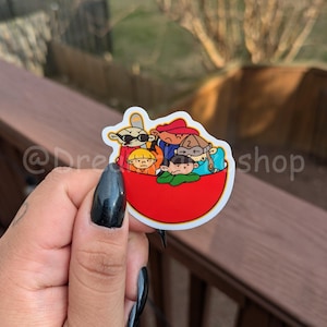 May include: A red bowl sticker featuring five cartoon characters. The characters are wearing different coloured clothing and hats. The sticker is on a wooden surface.