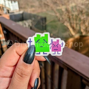 May include: A green and purple pixelated sticker of two characters. The green character has a sad expression and the purple character has an angry expression.
