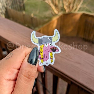 May include: A pink cartoon dog wearing a grey Viking helmet with horns and yellow braids. The dog has a worried expression on its face.