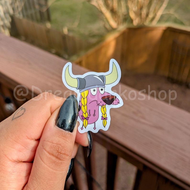 May include: A pink cartoon dog wearing a grey Viking helmet with horns and yellow braids. The dog has a worried expression on its face.