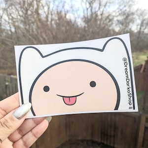 May include: White sticker with a cartoon face of Finn the Human from Adventure Time. Finn is smiling with his tongue sticking out. The sticker has a black outline and the text "@DreamDarkoShop" in the bottom right corner.