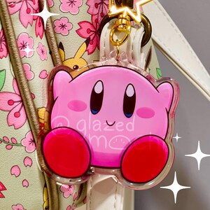 May include: A pink Kirby character keychain with red feet and the text "glazed moon". The keychain is attached to a gold-colored clasp with a star charm. The background includes a floral-patterned bag.