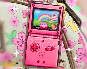 May include: A pink acrylic charm shaped like a retro handheld video game console. The screen displays a colorful scene with a rainbow, cake, and a character. The charm is attached to a keychain and is set against a floral background.