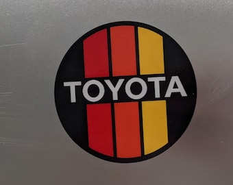 May include: A black and white circle sticker with red, orange, and yellow stripes. The word "TOYOTA" is written in white letters on the sticker.