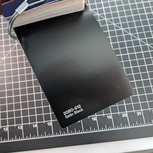 May include: A sample of 3M 2080-512 Satin Black vinyl wrap. The sample is on a black grid cutting mat.