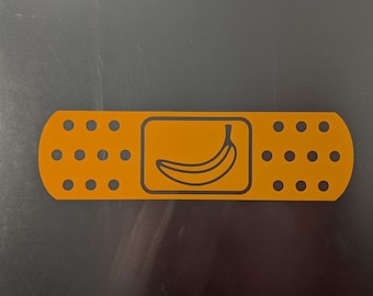 May include: A yellow band-aid with a black outline of a banana inside. The band-aid has a white border and a row of black dots on each side.