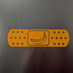Könnte beinhalten: Ein gelber Verband mit einer schwarzen Umrandung einer Banane im Inneren. Der Verband hat einen weißen Rand und eine Reihe schwarzer Punkte auf jeder Seite.