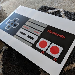 May include: A white poster with a black and gray Nintendo controller design. The controller has a directional pad, buttons labeled "A", "B", "Start", and "Select", and the Nintendo logo in red.
