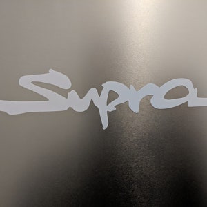 Puede incluir: Logotipo "Supra" blanco sobre un fondo gris. El logotipo está estilizado y escrito a mano.