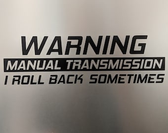 May include: A warning sign with black text on a white background that reads "WARNING MANUAL TRANSMISSION I ROLL BACK SOMETIMES."