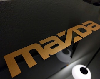May include: A gold-colored "MAZDA" logo on a black background.