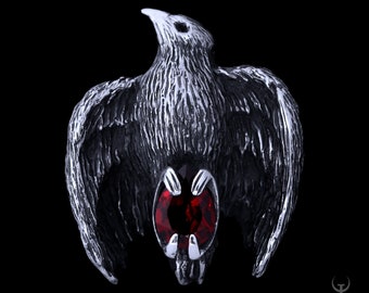 May include: A silver pendant in the shape of a crow with a red gemstone in its chest.