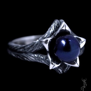 Op de afbeelding: Een zilveren ring met een donkerblauwe parel gezet in een bloemvormig ontwerp. De ringband is getextureerd en heeft een rustieke uitstraling.