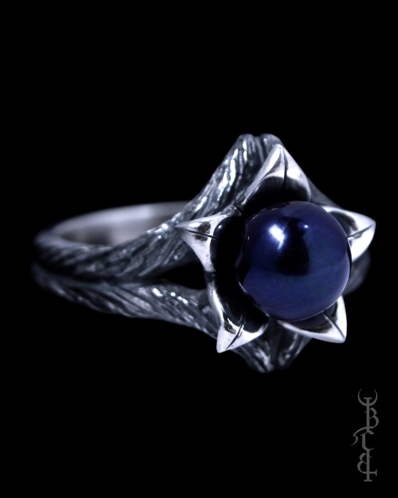 Op de afbeelding: Een zilveren ring met een donkerblauwe parel gezet in een bloemvormig ontwerp. De ringband is getextureerd en heeft een rustieke uitstraling.