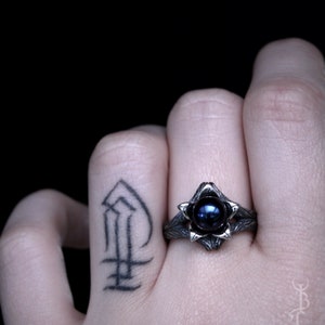 Op de afbeelding: Een zilveren ring met een donkerblauwe edelsteen gezet in een bloemvormig ontwerp. De ring wordt gedragen aan de vinger van een persoon met een tatoeage.
