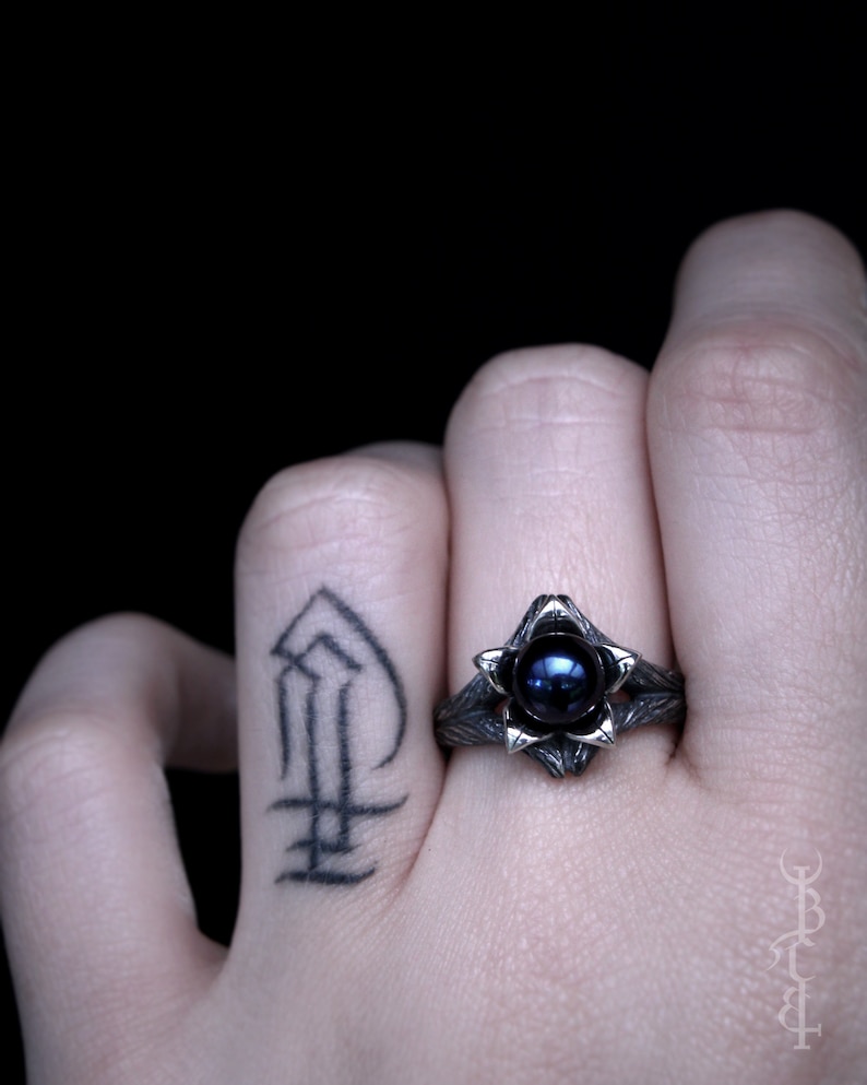 Op de afbeelding: Een zilveren ring met een donkerblauwe edelsteen gezet in een bloemvormig ontwerp. De ring wordt gedragen aan de vinger van een persoon met een tatoeage.