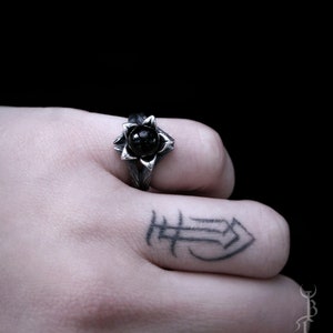 Op de afbeelding: Een zilveren ring met een zwarte edelsteen in het midden, in de vorm van een bloem. De ring wordt gedragen aan de vinger van een hand met een tatoeage.