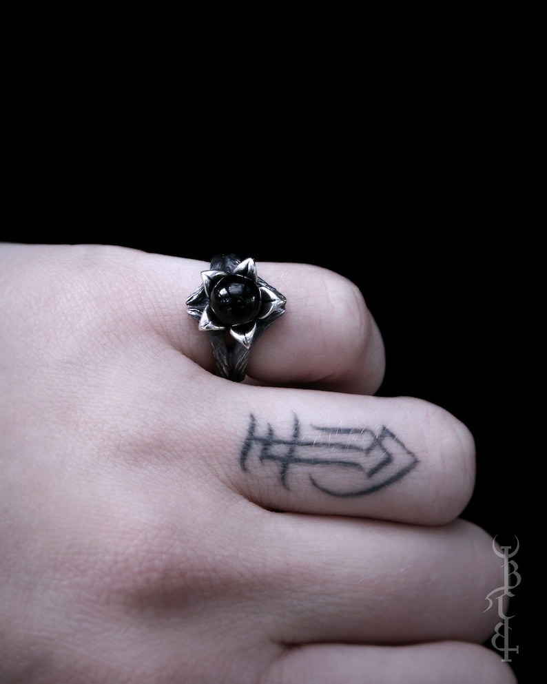 Op de afbeelding: Een zilveren ring met een zwarte edelsteen in het midden, in de vorm van een bloem. De ring wordt gedragen aan de vinger van een hand met een tatoeage.