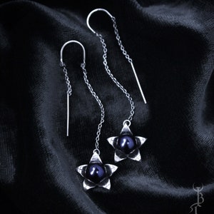 May include: A pair of silver dangle earrings with black pearl accents. Each earring features a five-pointed star design with a black pearl in the center.