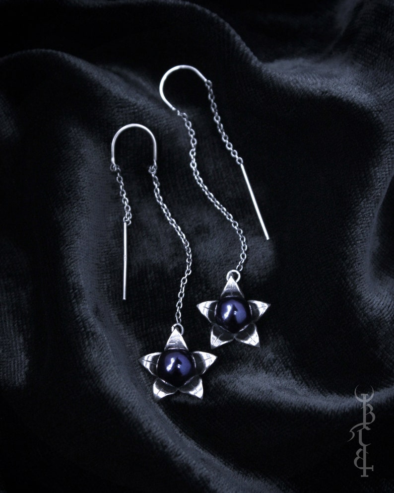 May include: A pair of silver dangle earrings with black pearl accents. Each earring features a five-pointed star design with a black pearl in the center.