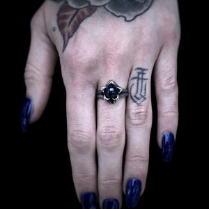 Op de afbeelding: Een zilveren ring met een zwarte edelsteen in het midden, omringd door een bloemmotief. De ring zit aan een vinger.