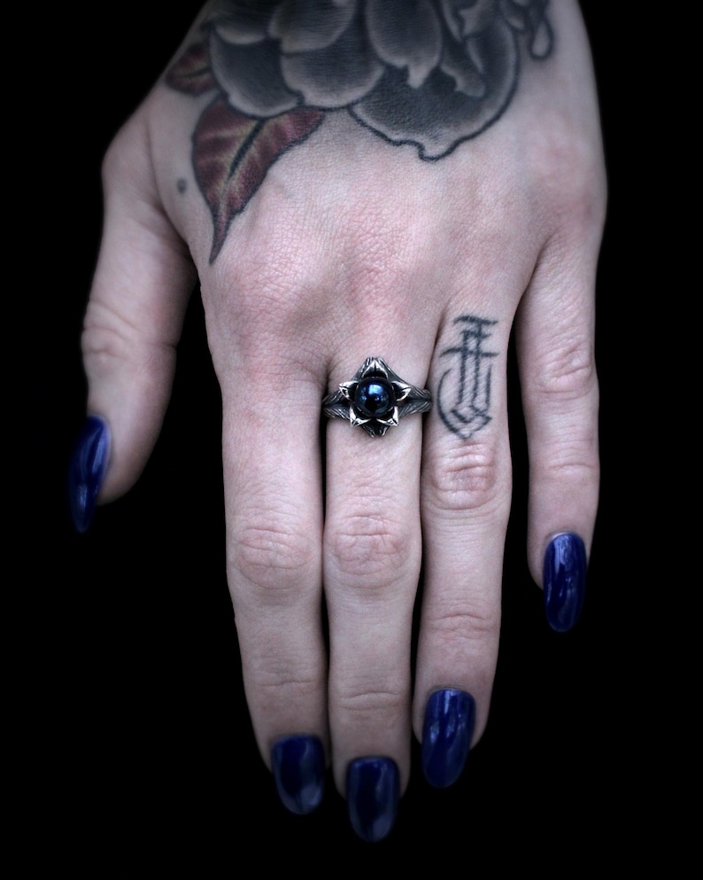 Op de afbeelding: Een zilveren ring met een zwarte edelsteen in het midden, omringd door een bloemmotief. De ring zit aan een vinger.