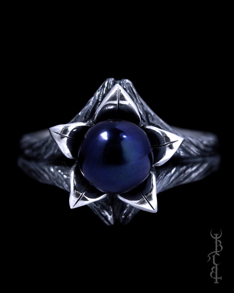 Op de afbeelding: Een zilveren ring met een donkerblauwe parel in een bloemvormig ontwerp.